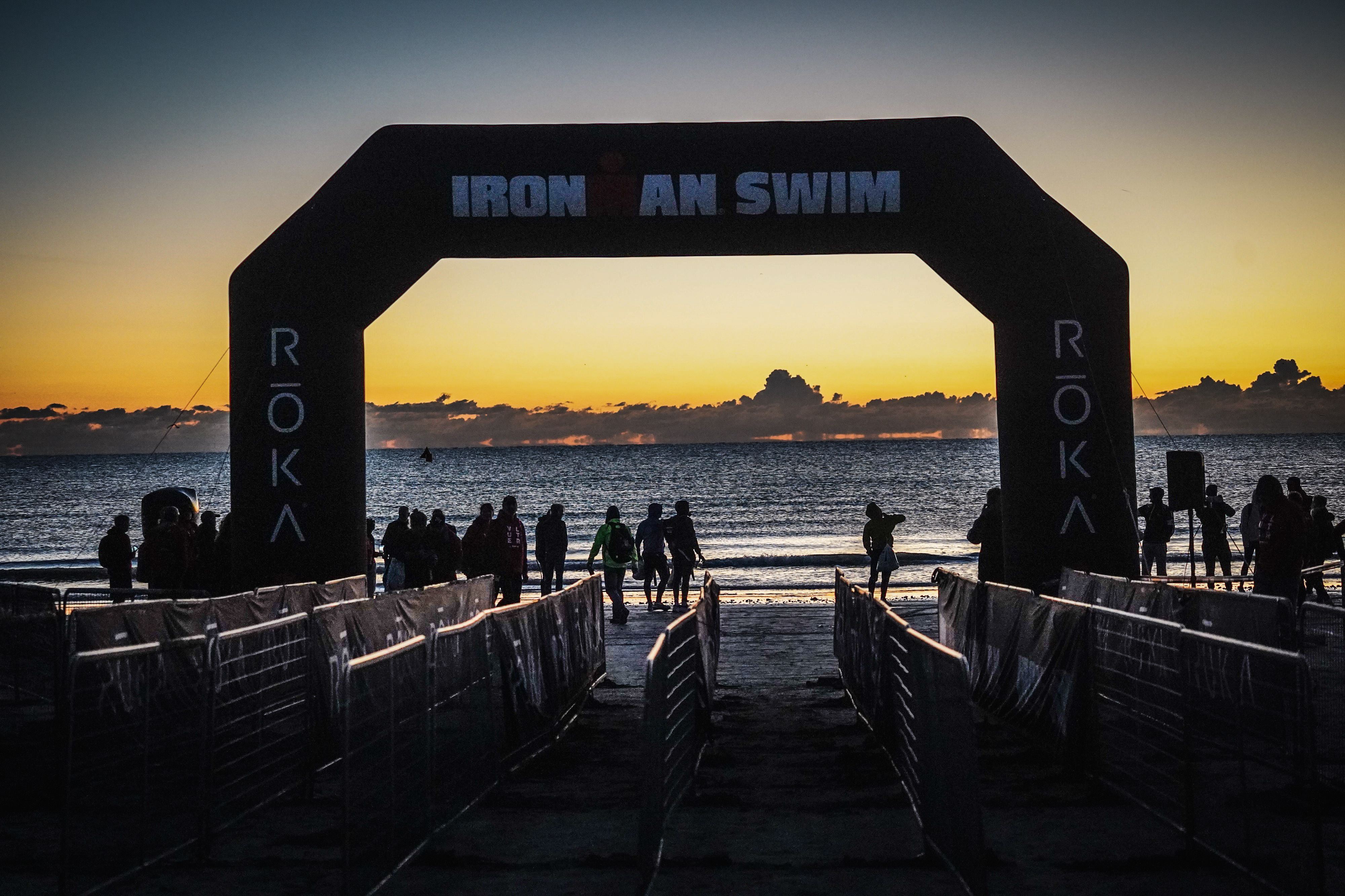 Sylwetki triatlonistów idących w stronę wody pod bramą 'IRONMAN SWIM' o wschodzie słońca, z żywym pomarańczowo-niebieskim niebem.