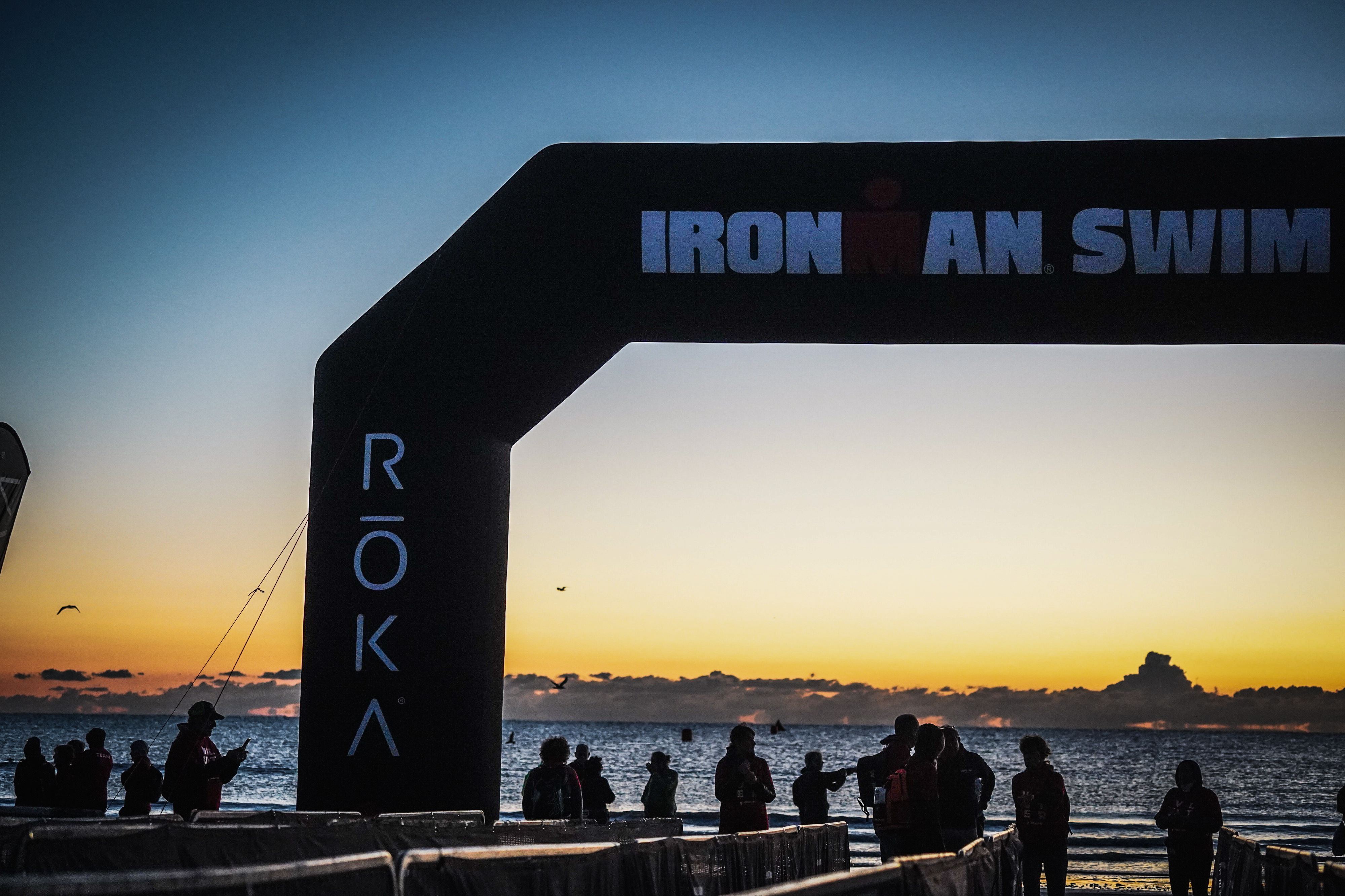 Sylwetki ludzi zgromadzonych w pobliżu bramy 'IRONMAN SWIM' na plaży o wschodzie słońca, z niebem w ciepłych odcieniach pomarańczu i błękitu.