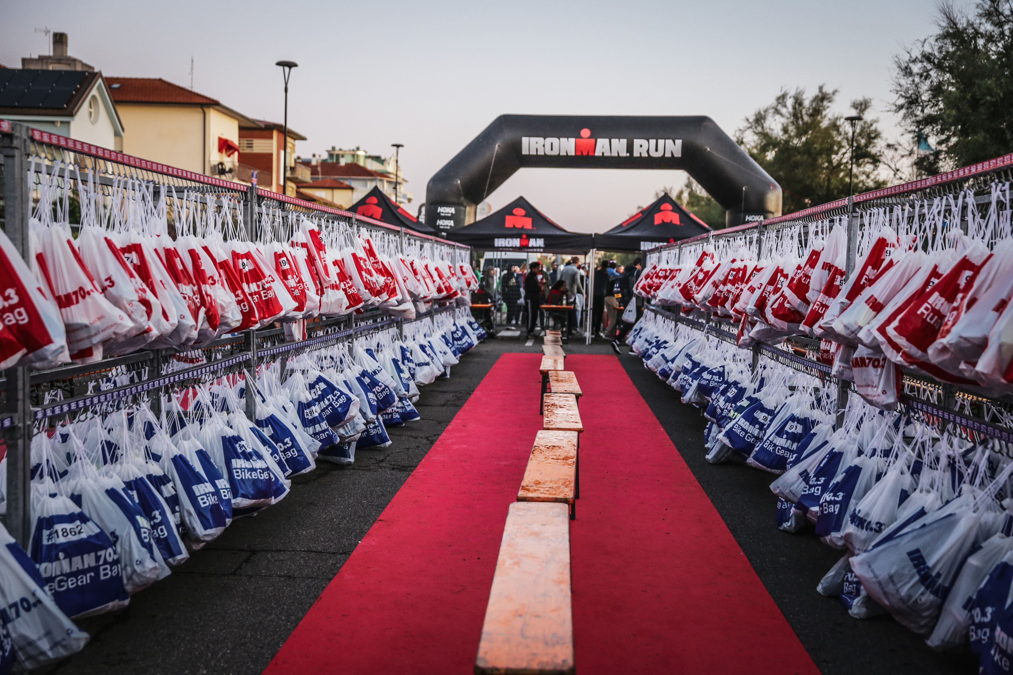 Długi czerwony dywan prowadzi do bramy 'IRONMAN RUN', otoczonej rzędami wiszących worków przejściowych (czerwono-białych, niebiesko-białych), gotowych na segment biegowy wyścigu.