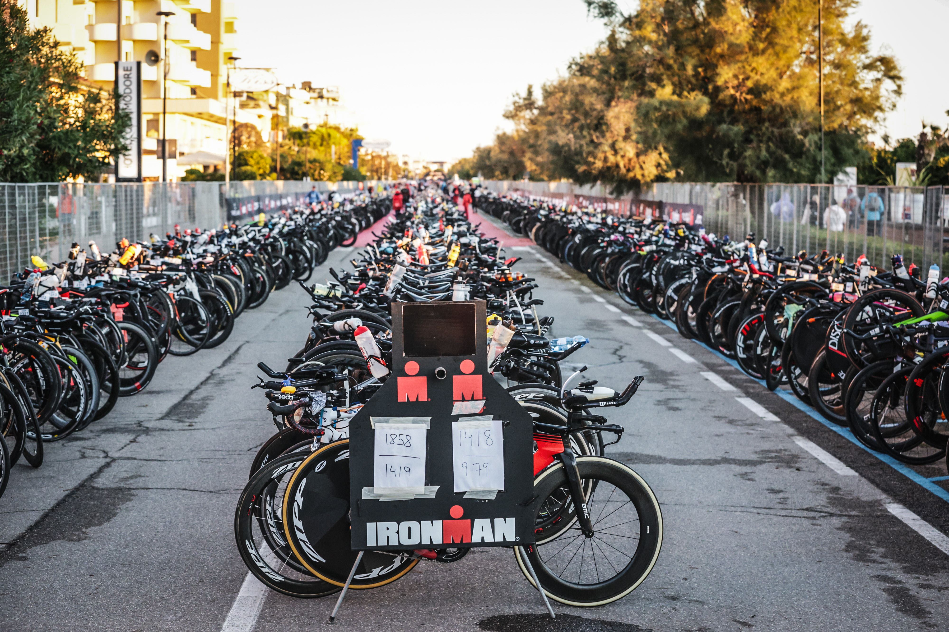 Rzędy rowerów są starannie zaparkowane w strefie zmian na imprezie Ironman, z dużym napisem 'IRONMAN' na pierwszym planie, wskazującym na początek segmentu kolarskiego.