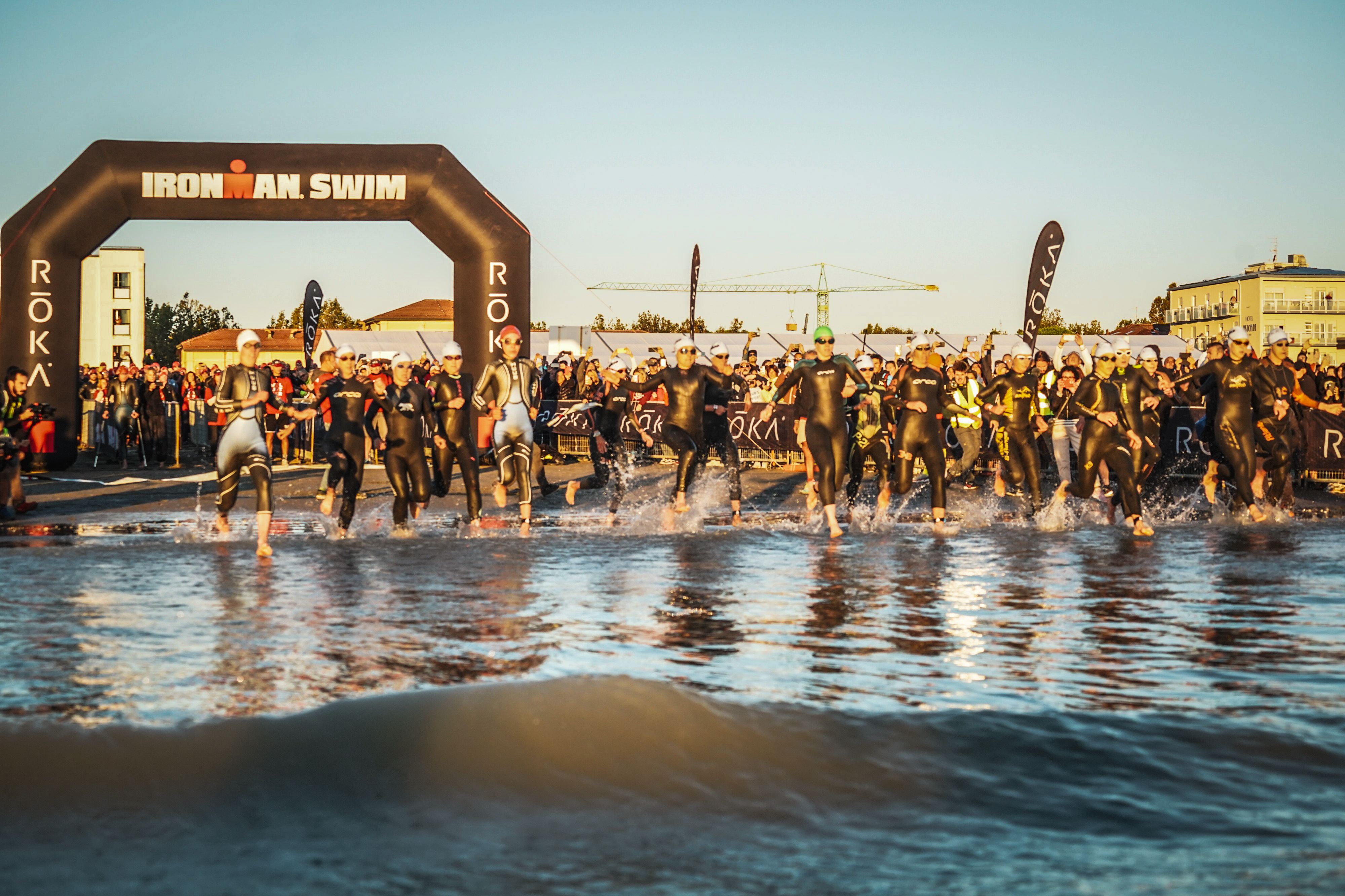 Triatloniści wbiegają do wody z plaży pod bramą 'IRONMAN SWIM', tworząc pluski, gdy rozpoczynają segment pływacki.