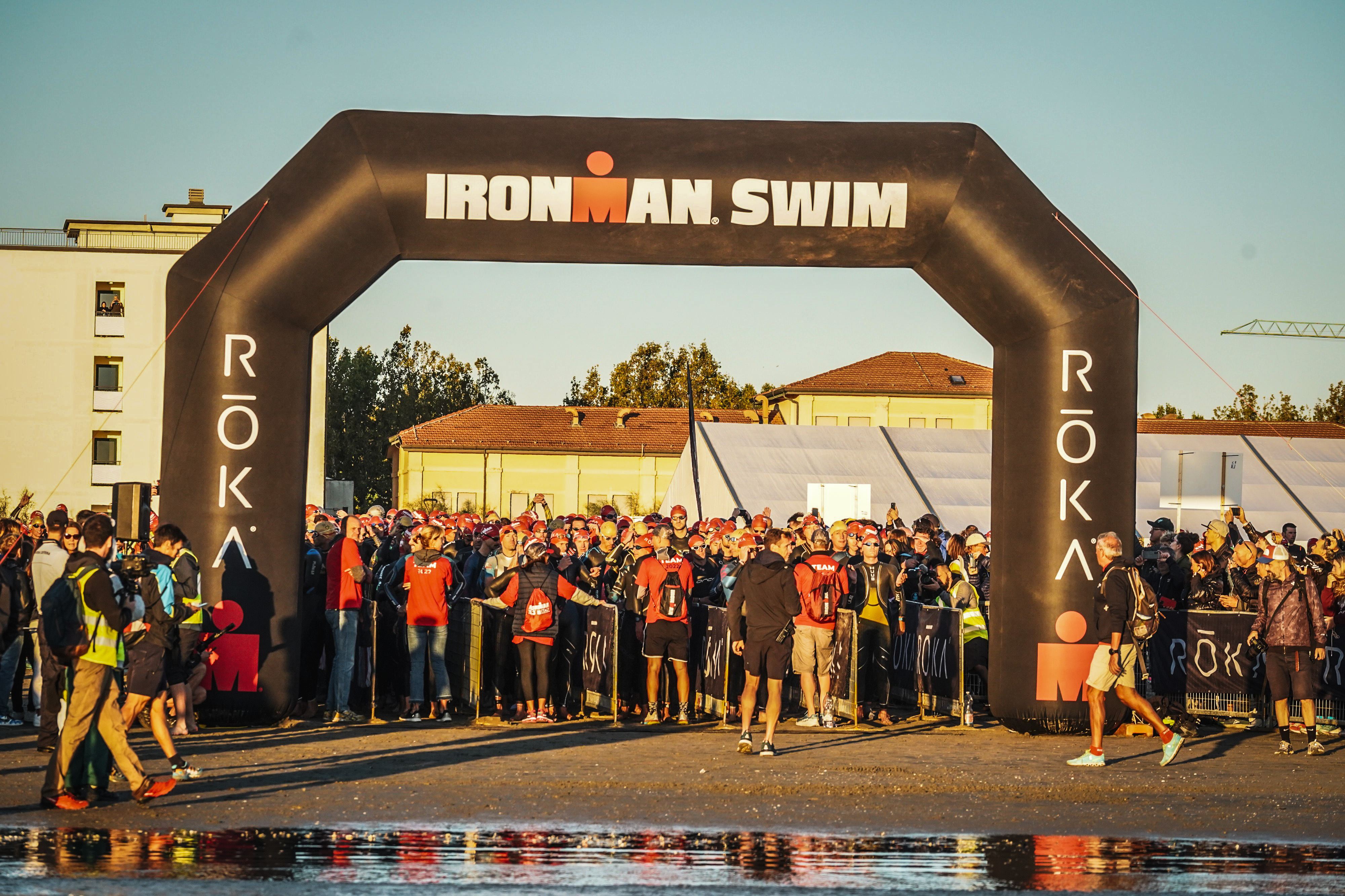Duża grupa triatlonistów w piankach zgromadzona pod bramą 'IRONMAN SWIM', przygotowująca się do startu pływackiego, z widzami w tle.