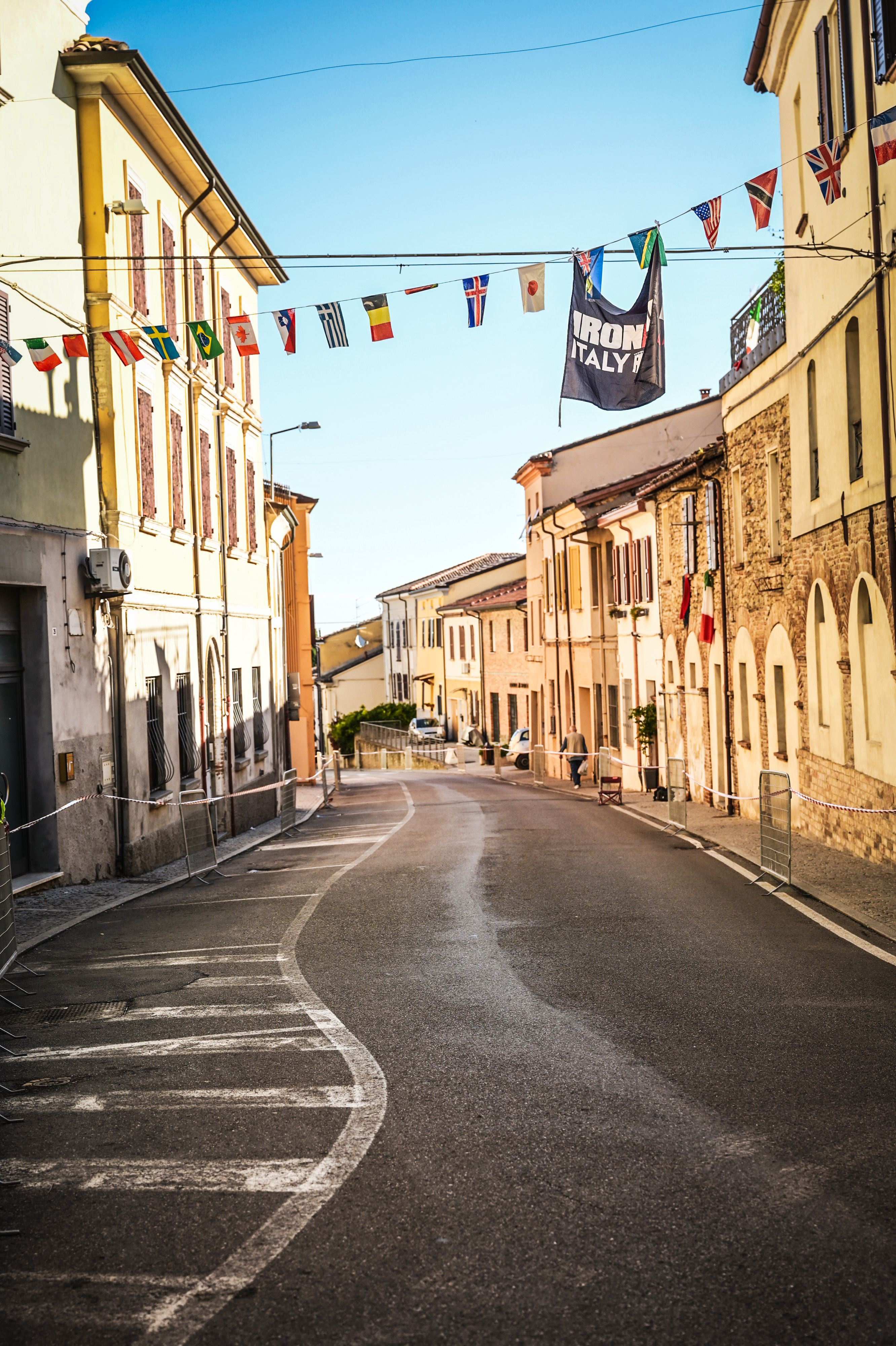 Wąska, kręta ulica w starym europejskim miasteczku, otoczona budynkami i ozdobiona flagami różnych krajów, z banerem 'IRONMAN ITALY' wiszącym nad głową.