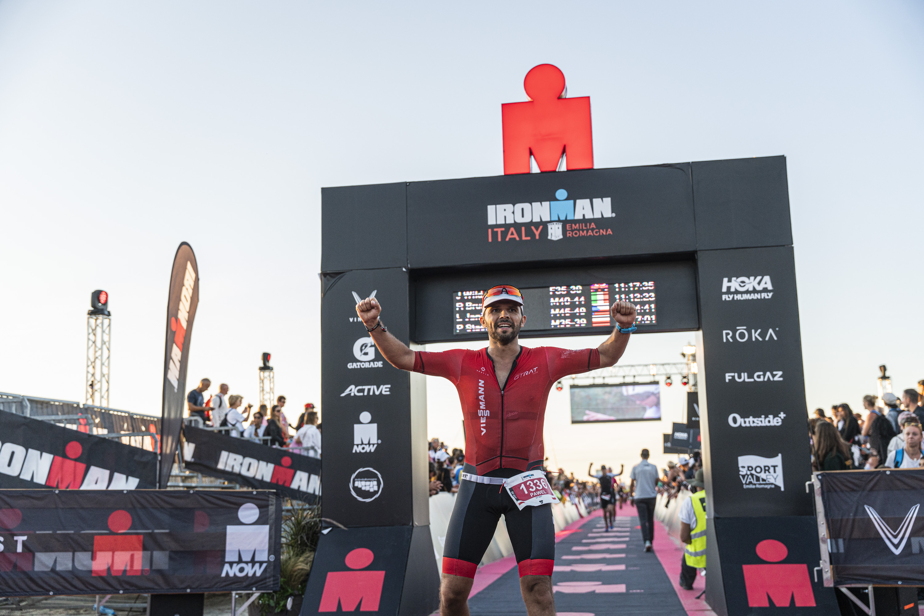 Mężczyzna triatlonista w czerwono-czarnym stroju stoi triumfalnie pod bramą mety Ironman, podnosząc ręce w geście zwycięstwa.